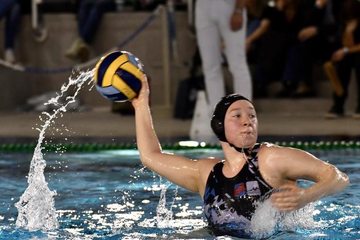 Wasserballspielerin wirft einen Ball im Sprung, während Wasser spritzt und Zuschauer im Hintergrund sitzen.