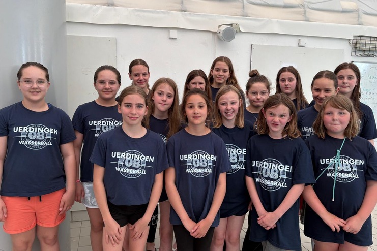 Gruppe von Mädchen in dunkelblauen T-Shirts mit „Uerdingen Wasserball“ Schriftzug, posierend in einer Sporthalle.