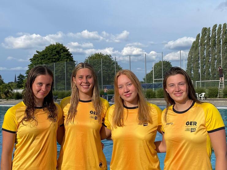 Vier junge Frauen in gelben Trikots posieren lächelnd am Rand eines Schwimmbeckens unter blauem Himmel.