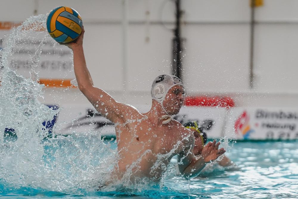 Wasserballspieler springt aus dem Wasser und wirft einen Ball in Richtung Tor, spritzendes Wasser um ihn herum.
