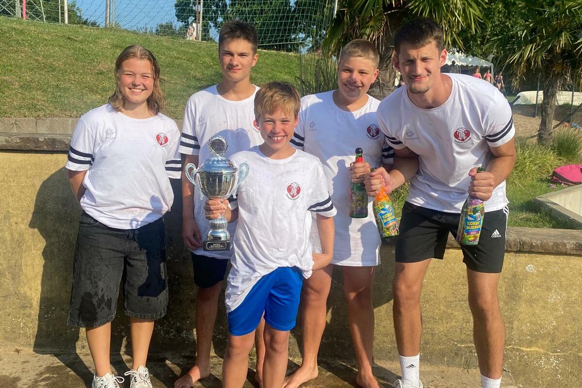 Fünf Kinder und Jugendliche stehen in Sportbekleidung mit Pokal und Getränken an einem Sportplatz.