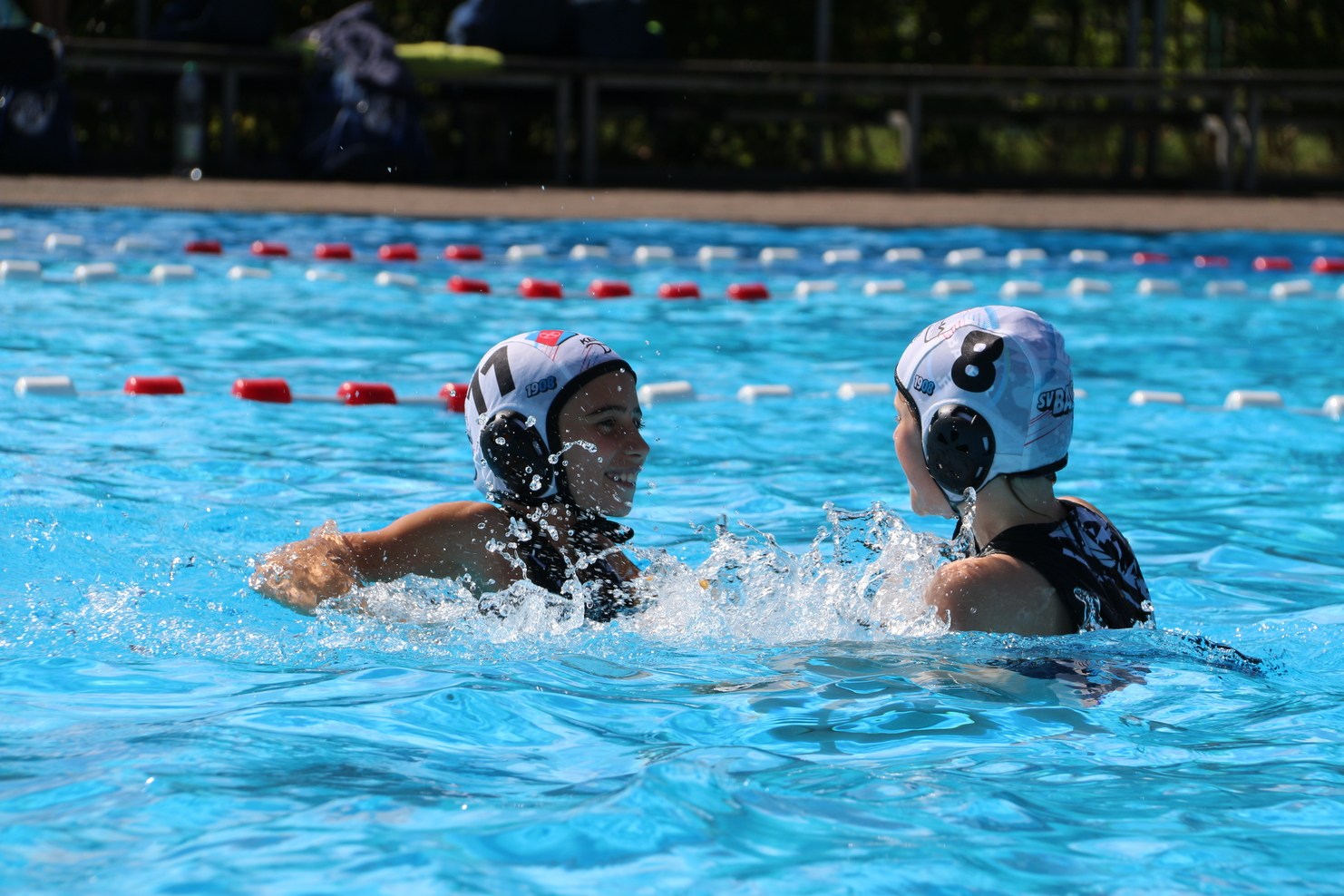 Zwei Wasserballspielerinnen lachen im splashtreibenden Wasser eines Schwimmbeckens mit roten und wei&szlig;en Markierungen.