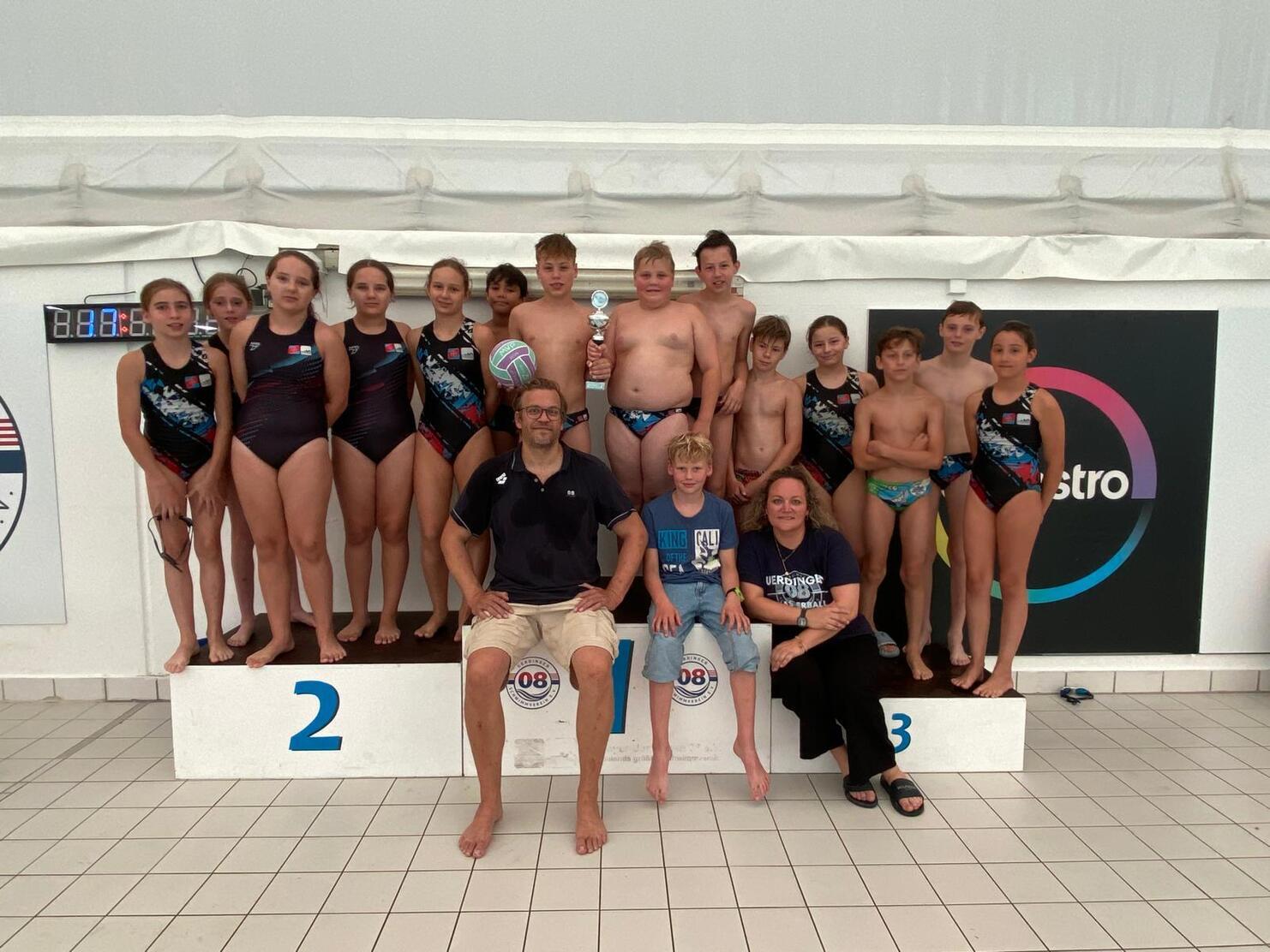 Gruppenfoto von Schwimmern in Badeanzügen auf dem Siegertreppchen mit Trainern und Pokal in einem Schwimmbad.