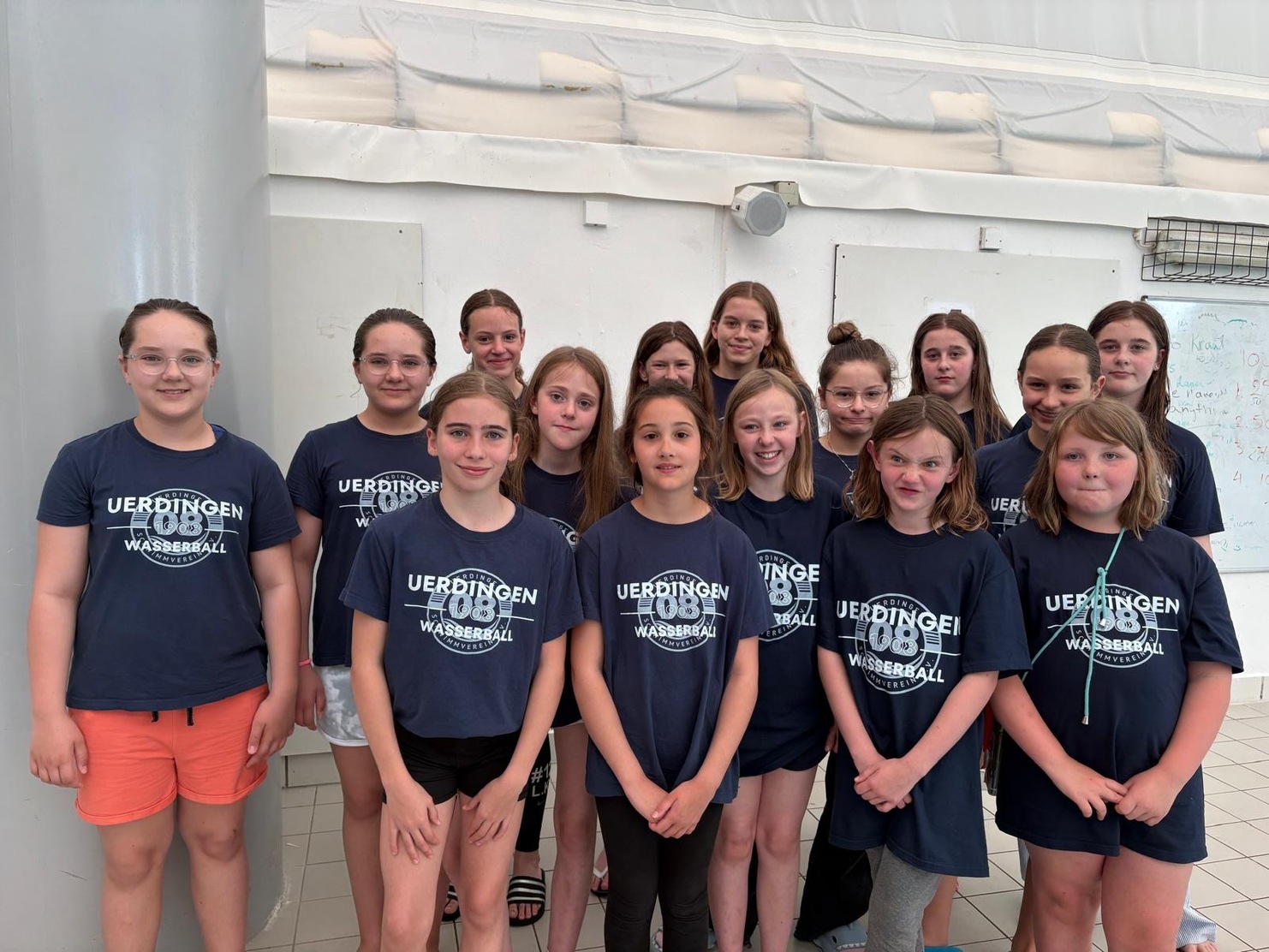 Gruppe von Mädchen in dunkelblauen T-Shirts mit „Uerdingen Wasserball“ Schriftzug, posierend in einer Sporthalle.