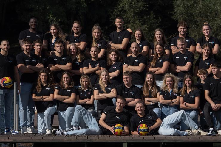 Gruppenaufnahme von 24 Personen in schwarzen T-Shirts, auf einer Holzplattform am Wasser, mit zwei Volleyballen.