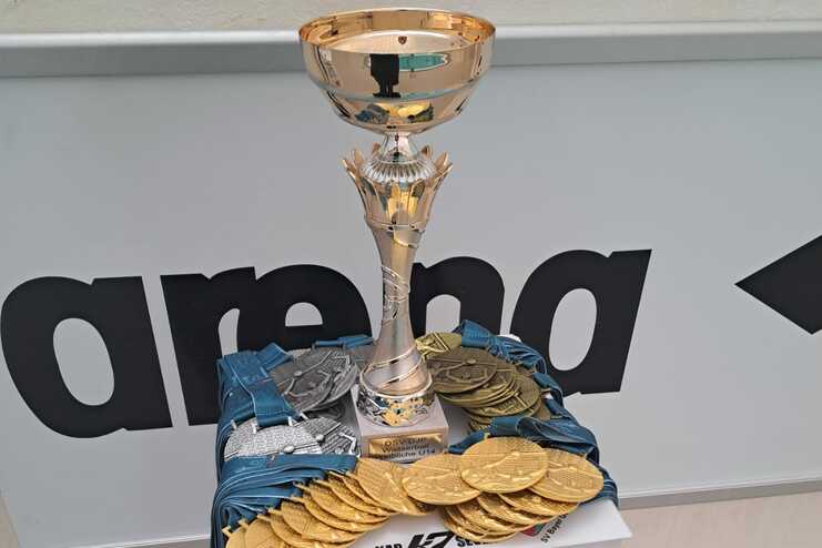 Silberner Pokal auf Podest mit vielen goldenen, silbernen und bronzenen Medaillen sowie blauen Bändern.