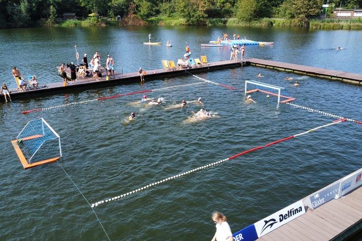 Aktive Schwimmer im Wasser auf einem Sportplatz mit Toren, während Zuschauer auf einem Steg entspannen.
