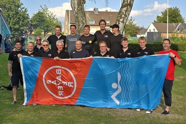Gruppenfoto von zehn Personen in Schwarz, die eine Flagge mit rotem und blauem Design halten, im Freien vor Bäumen.
