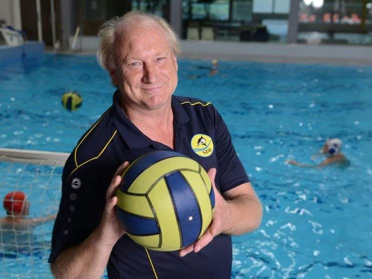 Wasserballtrainer hält einen Wasserball in der Hand, mit einem Schwimmbecken und weiteren Spielern im Hintergrund.