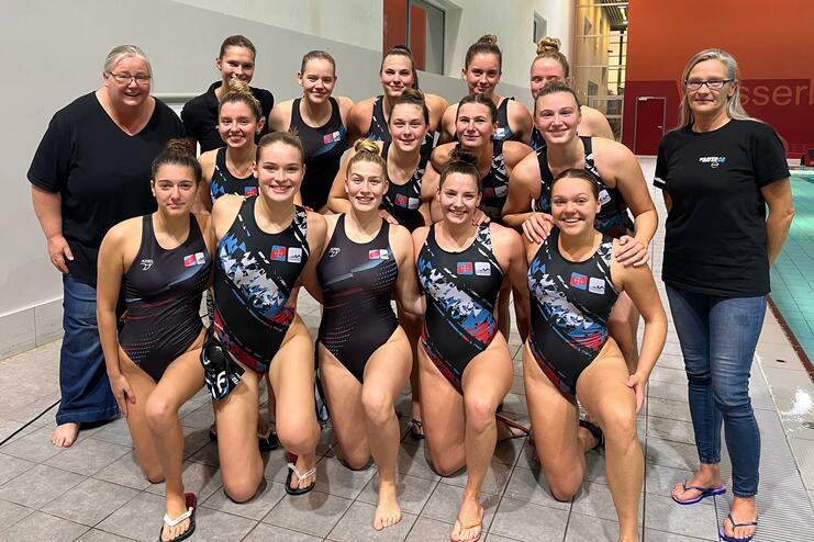 Gruppe von 14 Frauen im Wasserballanzug posiert lächelnd neben dem Pool, zwei Trainerinnen stehen hinter ihnen.
