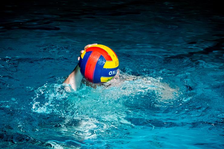 Wasserballspieler mit buntem Ball schwimmt im blauen Wasser eines Hallenbades.