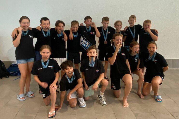 Gruppe von Kindern in schwarzen T-Shirts posiert mit Medaillen um den Hals in einem Schwimmbad.