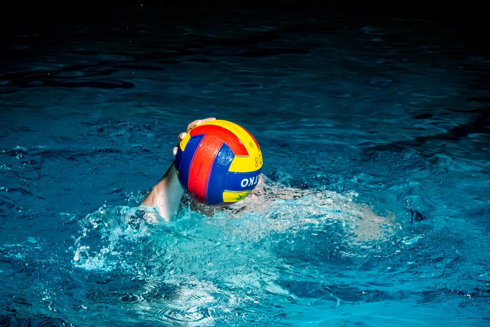 Wasserballspieler mit buntem Ball schwimmt im blauen Wasser eines Hallenbades.