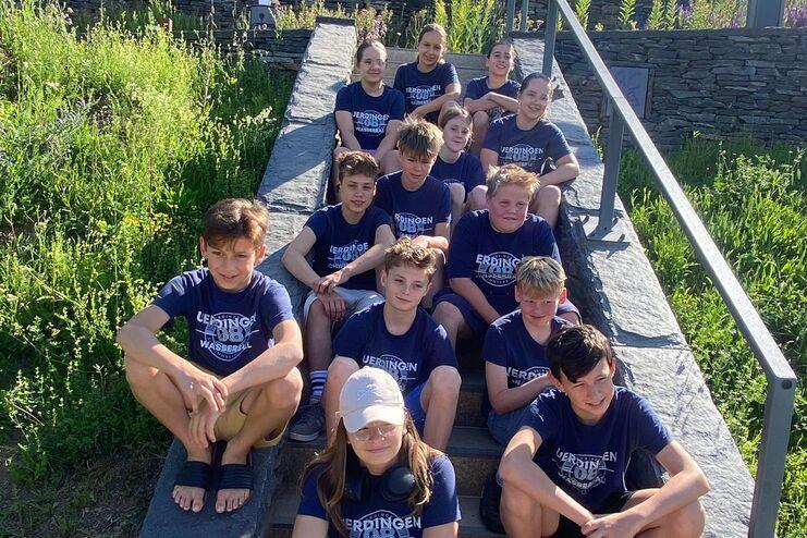 Gruppe von Kindern in blauen T-Shirts sitzt auf Treppe, umgeben von grüner Landschaft und blühenden Pflanzen.