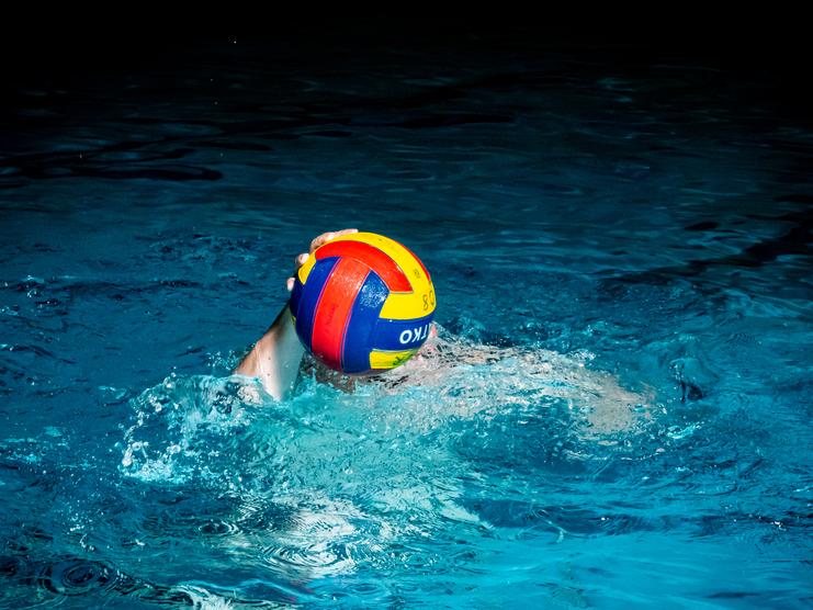 Wasserballspieler mit buntem Ball schwimmt im blauen Wasser eines Hallenbades.