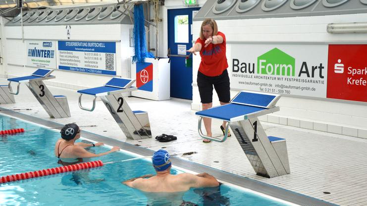 Trainerin leitet Schwimmer am Beckenrand eines Schwimmbads mit Startblöcken und Werbebannern.
