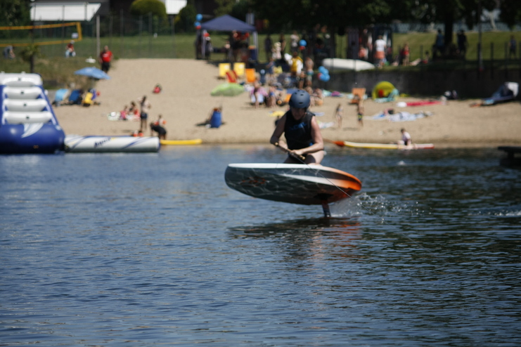 Junge fährt auf einem Foil-Board über das Wasser, im Hintergrund sichtbare Badelandschaft und Strandgäste.