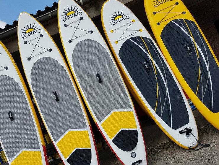 Fünf Stand-Up-Paddle-Boards in verschiedenen Farben stehen in Reihe auf einem Pflasterboden.