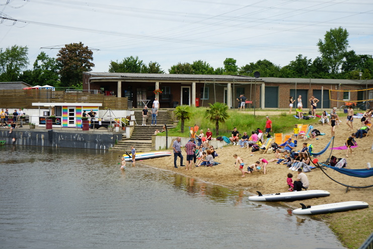Badegäste entspannen am Ufer eines Sees, umgeben von Sand, Liegen und Wassersportgeräten.