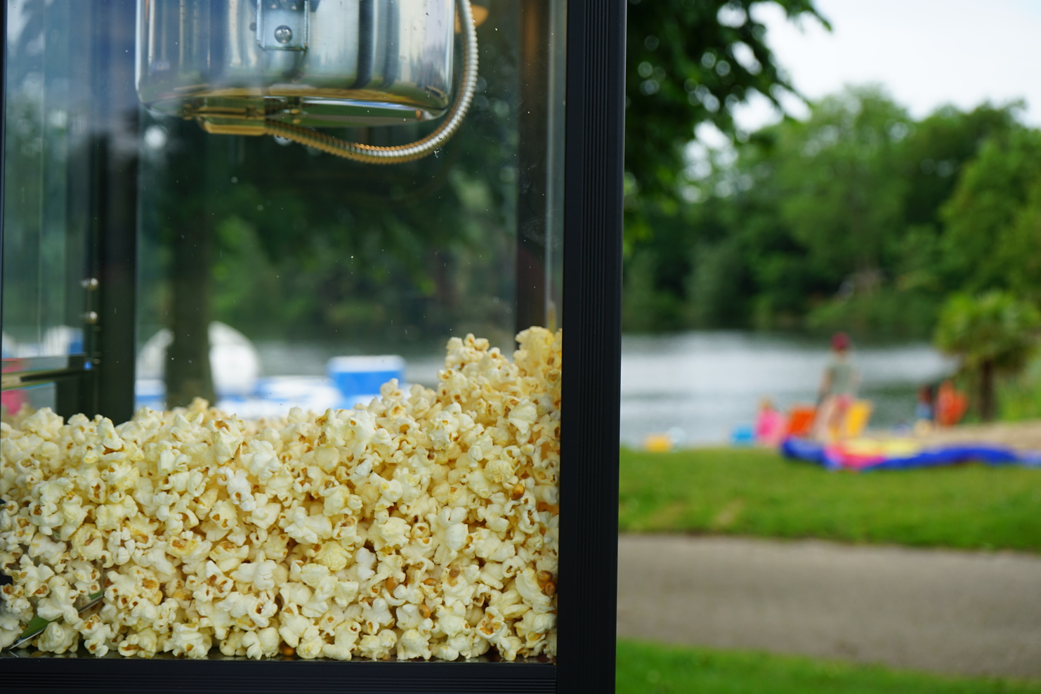 Nahaufnahme einer Popcornmaschine mit frischem Popcorn, im Hintergrund ein See und unscharfe Personen am Ufer.