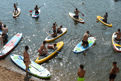 Verschiedene Personen paddeln auf Stand-Up-Paddle-Boards auf einem Gewässer, umgeben von einer sandigen Uferpromenade.