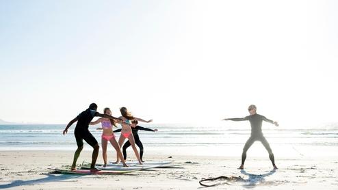 Fünf Personen in Neoprenanzügen üben Surfen auf Surfbrettern am Strand bei sonnigem Wetter.