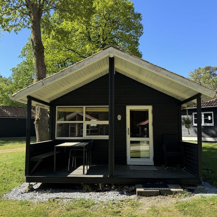 Schwarzes Ferienhaus mit Veranda, Tisch und St&uuml;hlen vor einer gr&uuml;nen Wiese, umgeben von B&auml;umen und blauem Himmel.