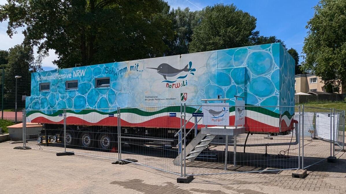Rollende Schwimmstation mit buntem Design, umgeben von einem Zaun, steht auf einem Platz mit Bäumen im Hintergrund.