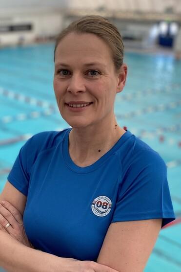 Eine Frau in blauem Sportshirt steht lächelnd mit verschränkten Armen vor einem Schwimmbecken.