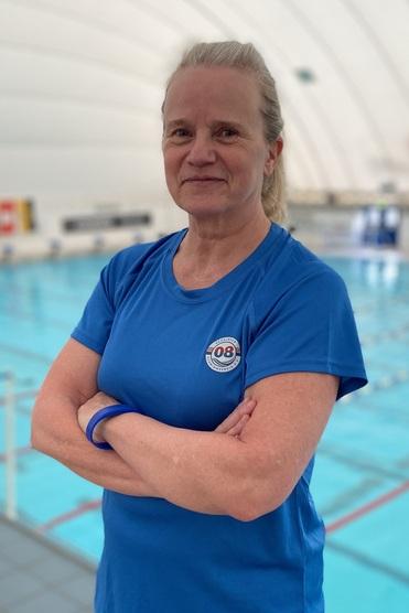 Frau in blauer Sportbekleidung steht am Beckenrand eines Schwimmbeckens mit blauer Wasseroberfläche im Hintergrund.