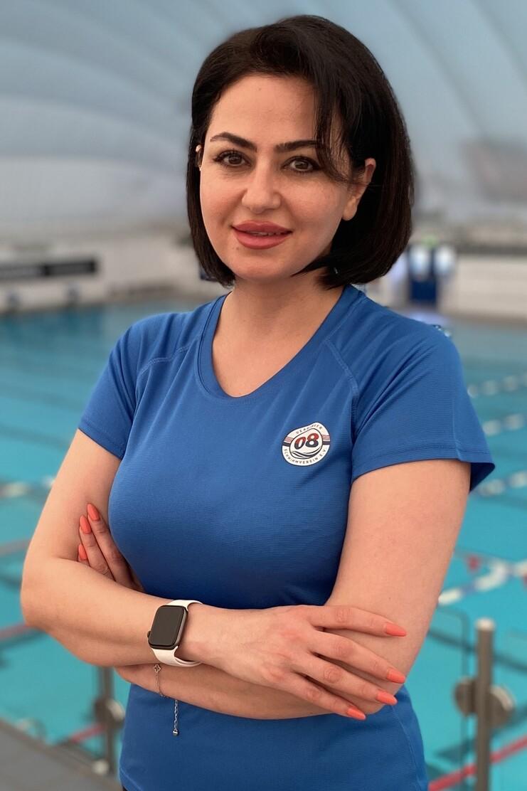 Frau in blauer Sportbekleidung steht mit verschr&auml;nkten Armen vor einem Schwimmbecken in einer Sporthalle.