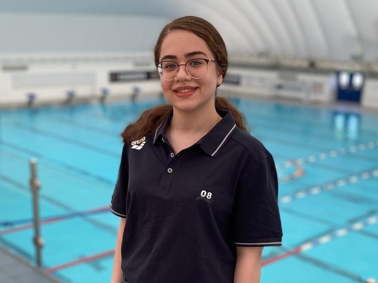 Junge Frau in Freizeitkleidung steht am Rand eines Schwimmbeckens in einer Halle mit Sportanlagen.