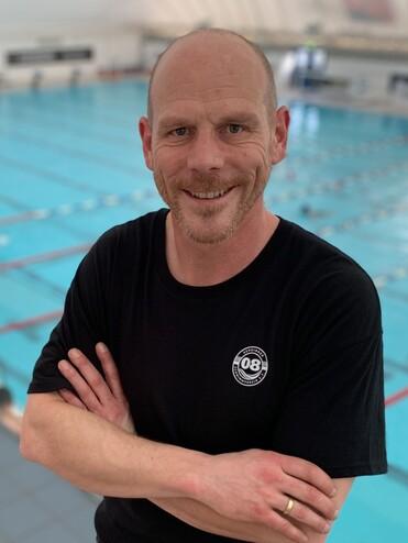 Mann mit kurzen Haaren in schwarzem T-Shirt steht mit verschr&auml;nkten Armen vor einem Schwimmbecken.