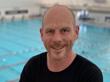 Mann mit kurzen Haaren in schwarzem T-Shirt steht mit verschr&auml;nkten Armen vor einem Schwimmbecken.