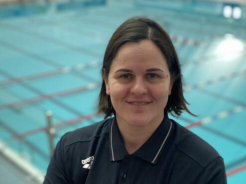 Frau mit kurzem Haar in Sportkleidung steht vor einem leeren Schwimmbecken, lächelt und hat die Arme vor der Brust verschränkt.