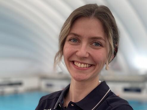Lächelnde Frau im blauen Polohemd steht mit verschränkten Armen an einem Schwimmbecken.