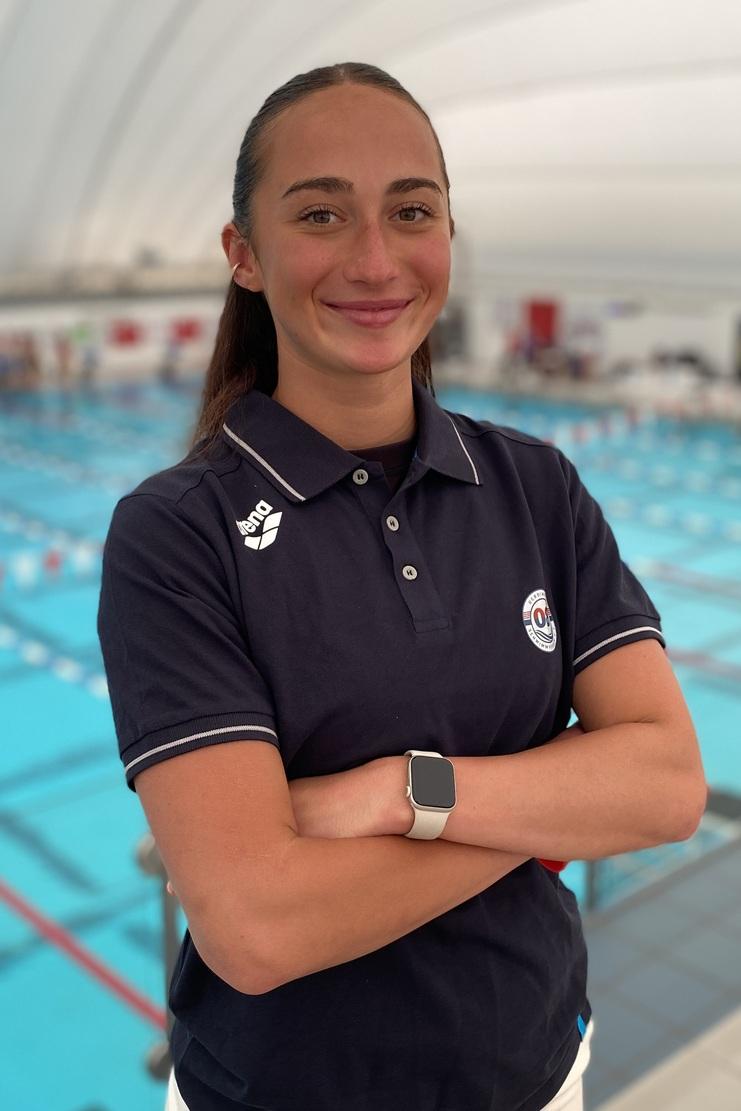 Lächelnde Trainerin in blauer Sportkleidung steht am Beckenrand eines Schwimmbads mit blauen Wasserbahnen im Hintergrund.