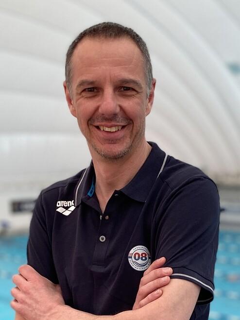 Mann mit kurzem Haar, in schwarzem Polo-Shirt, lächelt vor blauer Schwimmbeckenlandschaft in einer Schwimmhalle.