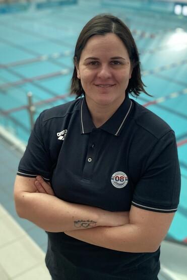 Frau mit kurzem Haar in Sportkleidung steht vor einem leeren Schwimmbecken, l&auml;chelt und hat die Arme vor der Brust verschr&auml;nkt.