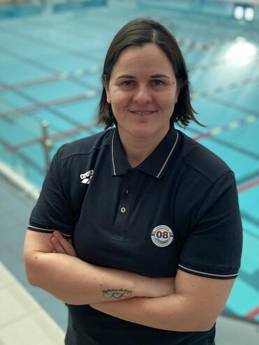 Frau mit kurzem Haar in Sportkleidung steht vor einem leeren Schwimmbecken, l&auml;chelt und hat die Arme vor der Brust verschr&auml;nkt.