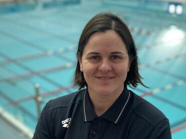 Frau mit kurzem Haar in Sportkleidung steht vor einem leeren Schwimmbecken, l&auml;chelt und hat die Arme vor der Brust verschr&auml;nkt.