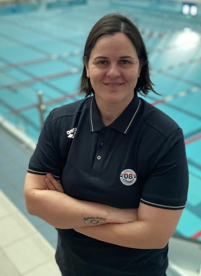 Frau mit kurzem Haar in Sportkleidung steht vor einem leeren Schwimmbecken, lächelt und hat die Arme vor der Brust verschränkt.