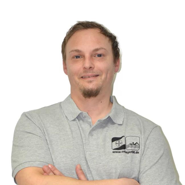 Lächelnder Mann mit kurzem Haar und Bart trägt ein graues Poloshirt und hat die Arme vor der Brust verschränkt.