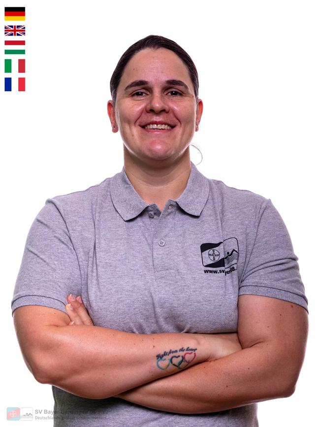 Eine lächelnde Frau mit kurzen Haaren trägt ein graues Polohemd und hat die Arme vor der Brust verschränkt.