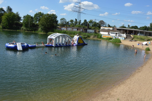 Blick auf einen See mit Schwimmern und einem aufblasbaren Spielgerät, umgeben von Sträuchern und Sandstrand.
