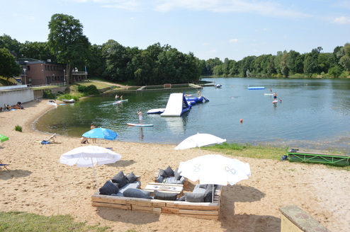 Strandbereich an einem See mit Liegen, Sonnenschirmen, einem Wasserspielplatz und Leuten beim Stand-Up-Paddling.