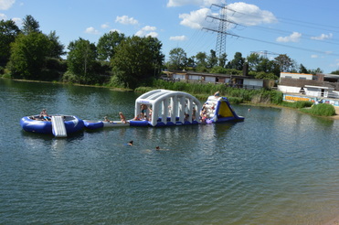 Blick auf einen See mit Schwimmern und einem aufblasbaren Spielgerät, umgeben von Sträuchern und Sandstrand.