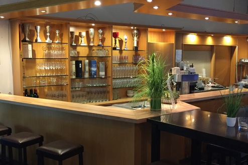 Theke mit Gläsern, Pokalen und Pflanzen in einem modernen Barbereich. Sitzgelegenheiten aus Holz sind darauf ausgerichtet.