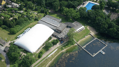 Luftaufnahme eines Freizeitgeländes mit Sporthalle, Schwimmbad und Anlegestelle am glitzernden Wasser.