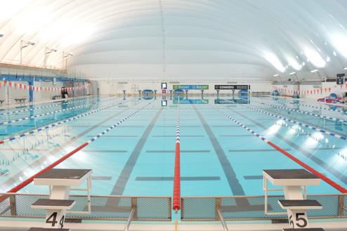 Innenansicht eines beleuchteten Schwimmbads mit mehreren Bahnen, Startbl&ouml;cken und bunten Wendenflaggen.
